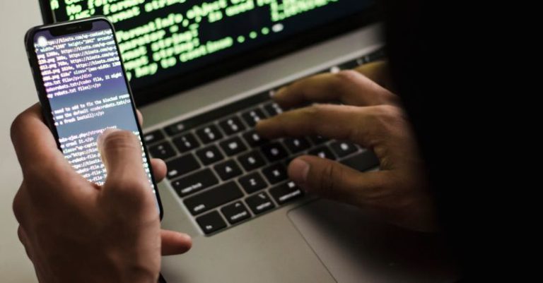 Access Control - Unrecognizable hacker with smartphone typing on laptop at desk